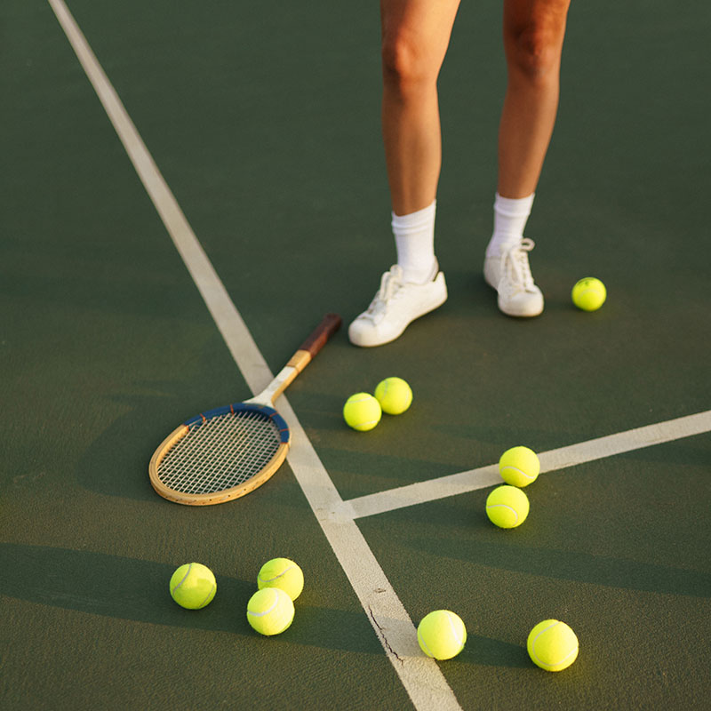 En person som lagt sitt tennisrack på banan med flera tennisbollar vid sidan.